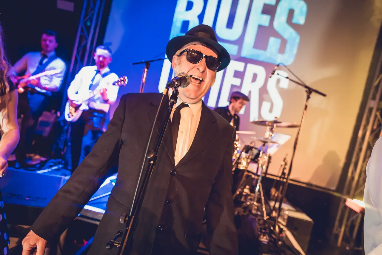 The Blues Others' frontman in a black suit, trilby and shades singing on stage